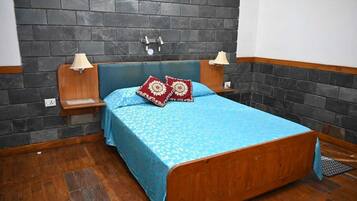 A bedroom with a wooden bed, a blue patterned bedspread, two decorative pillows, a nightstand with a lamp, and a wall with a black and grey tile pattern.
