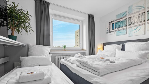 Una habitació d'hotel amb un llit gran, un altre més petit, una planta en test, una finestra amb cortines i obra d'art a la paret.