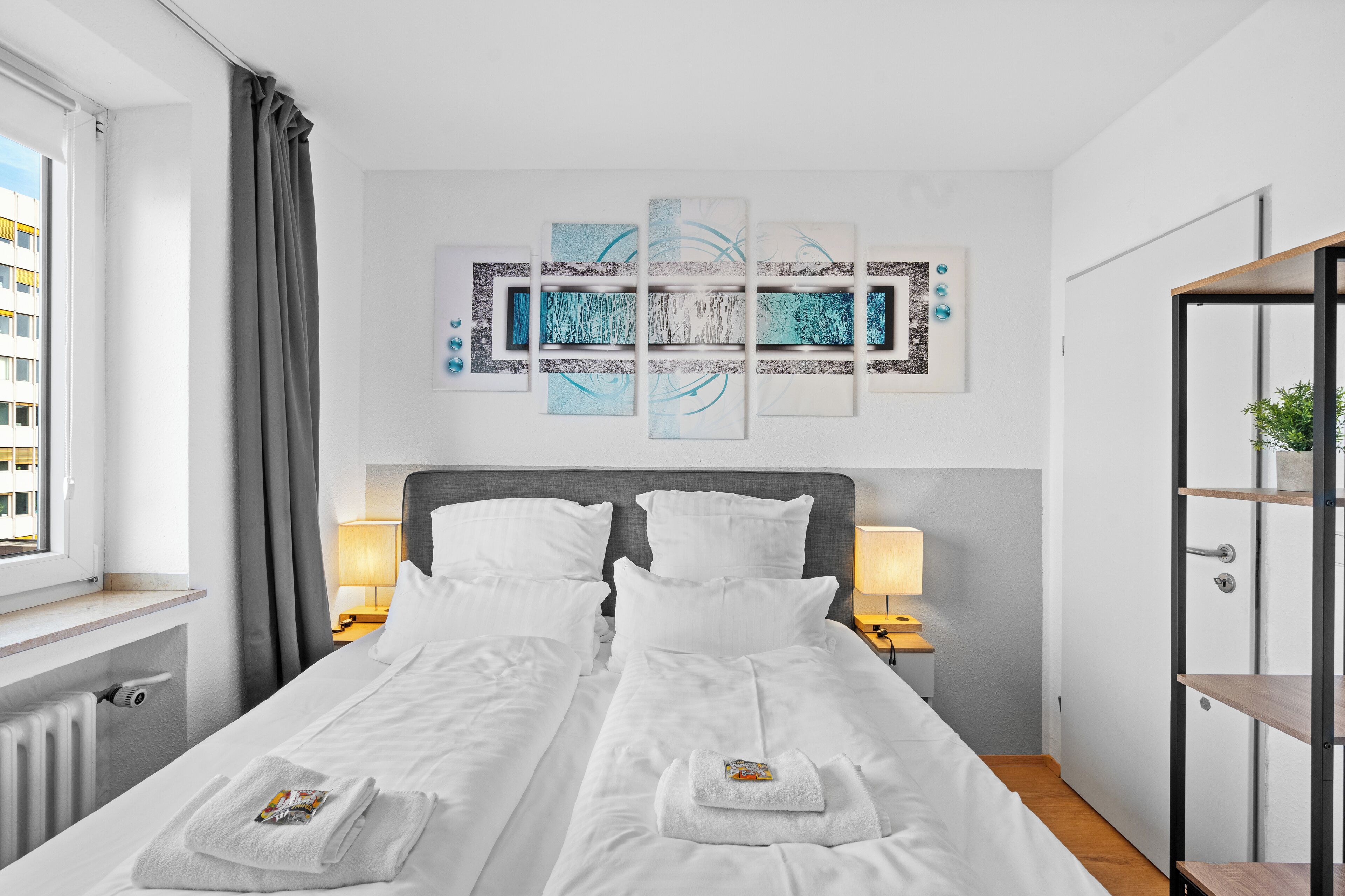 Une chambre d’hôtel avec un grand lit, deux tables de chevet avec des lampes, une œuvre d’art murale et une vue sur un bâtiment à l’extérieur.