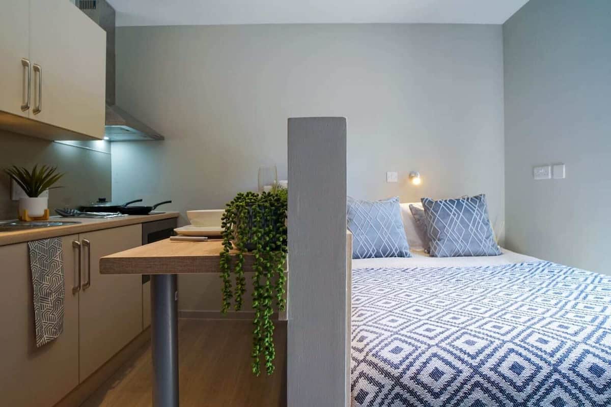 Una habitación pequeña con kitchenette, una cama con ropa de cama estampada y una planta en maceta.