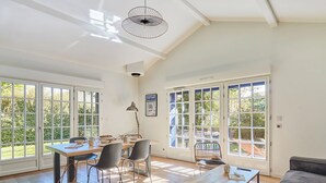 A spacious room with a wooden floor, a dining table with chairs, a sofa, and large windows offering a view of the outdoors. - Villas Odalys Collection Domaine de Lana by Interhome (Saint-Jean-de-Luz)
