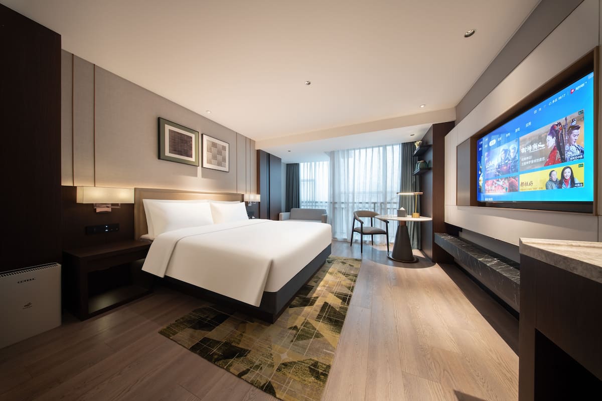 A modern hotel room with a large bed, a flat-screen TV, a desk, and a view of the city through large windows.