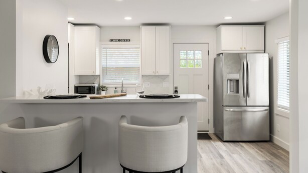 A modern kitchen with white cabinetry, a stainless steel refrigerator, and a bar area with two white barstools.