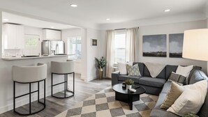 A modern living room with a grey sofa, two bar stools, a coffee table, and a kitchen area with white cabinets and a refrigerator.