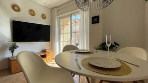 Un coin repas avec une table ronde, des chaises blanches, un téléviseur à écran plat fixé au mur et une fenêtre avec des rideaux.