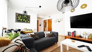 Un salon moderne avec un canapé gris, une table basse blanche et un téléviseur à écran plat. On y trouve des coussins décoratifs, une plante et un lustre noir original.