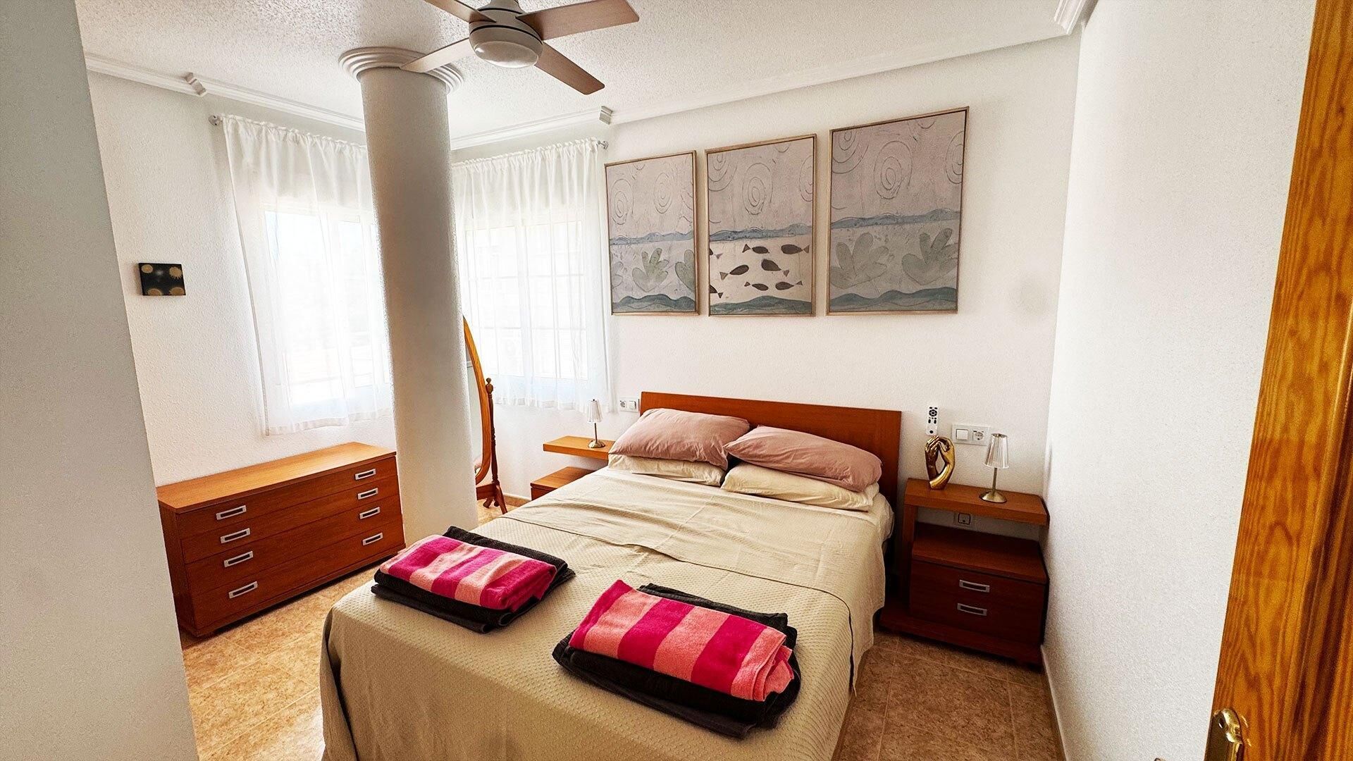 A bedroom with a bed, bedside tables, a dresser, a ceiling fan, and artwork on the wall.