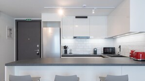 A modern kitchen with white cabinets, a stainless steel refrigerator, a microwave, and a red kettle. There are three grey bar stools in front of the counter. - LIV Athinum 1 - Luxury Apartment 3B (Athens)