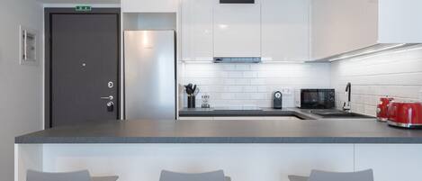 A modern kitchen with white cabinets, a stainless steel refrigerator, a microwave, and a red kettle. There are three grey bar stools in front of the counter.