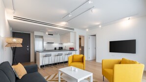 A modern living room with a sofa, two yellow armchairs, a white coffee table, and a kitchen in the background. - LIV Athinum 1 - Luxury Apartment 3B (Athens)