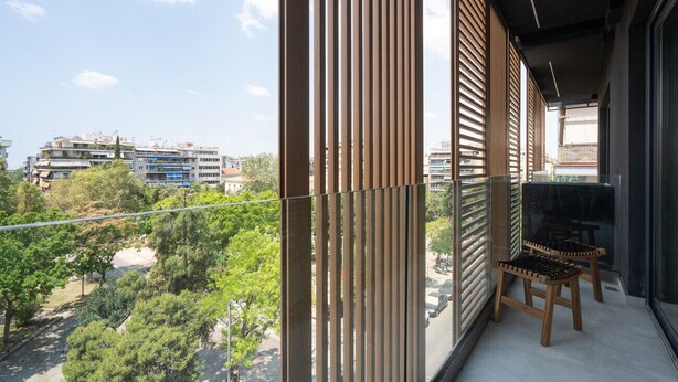 A balcony with vertical blinds, a pair of wooden chairs, and a view of a cityscape with buildings and trees. - LIV Athinum 1 - Luxury Apartment 3B (Athens)