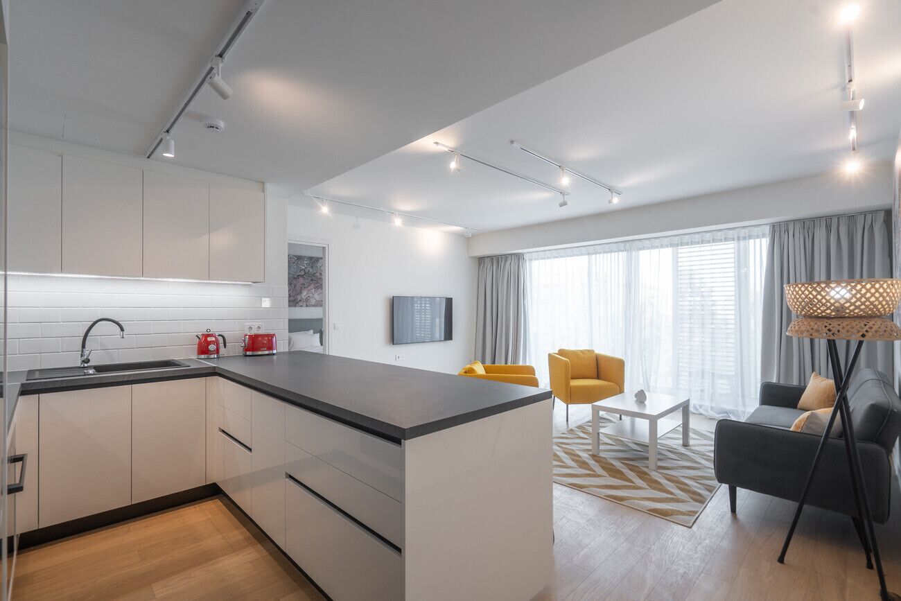 Eine moderne Küche mit weißen Schränken, einer schwarzen Arbeitsplatte und einem Spülbecken. Es gibt eine Essecke mit einem gelben Stuhl und einem grauen Sofa.
