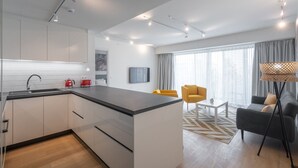 A modern kitchen with white cabinets, a black countertop, and a sink. There is a dining area with a yellow chair and a grey sofa. - LIV Athinum 1 - Luxury Apartment 3B (Athens)