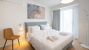 A modern bedroom with a bed, a chair, a painting, and a window with curtains. - LIV Athinum 1 - Luxury Apartment 3B (Athens)