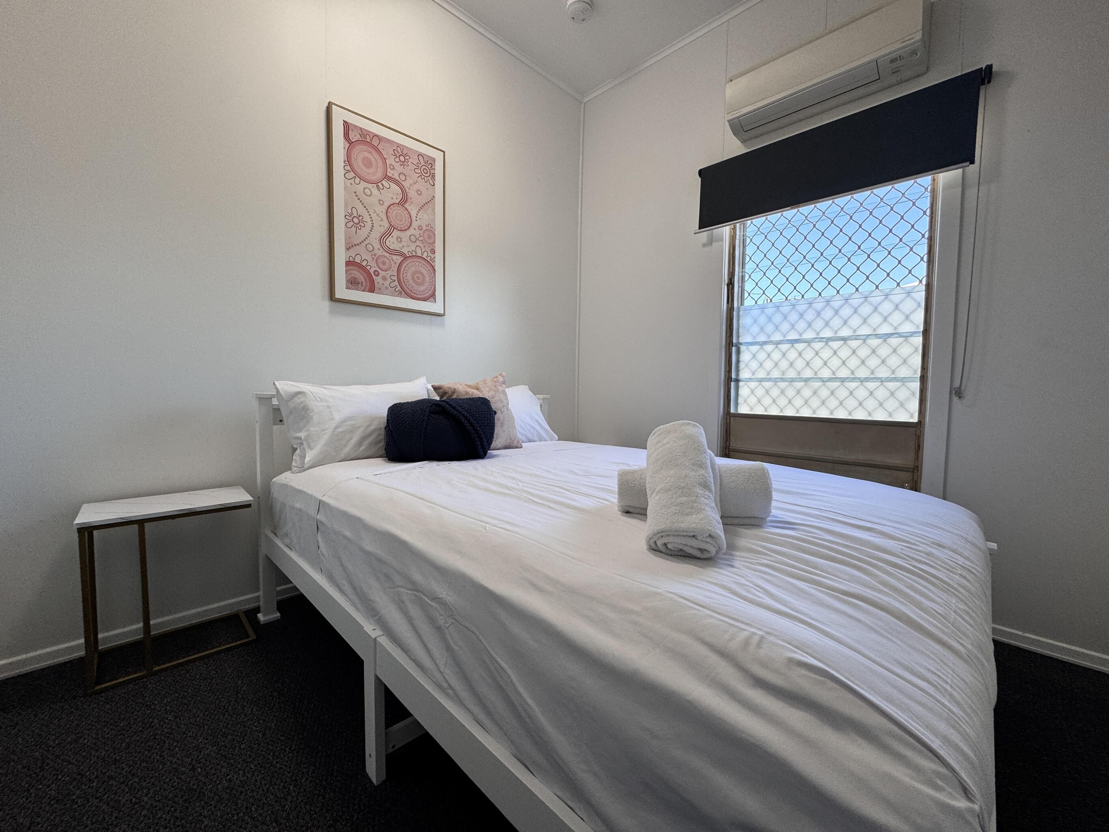 A neatly made bed with white linens and two rolled towels, a small bedside table, a framed artwork on the wall, and a window with a metal screen.
