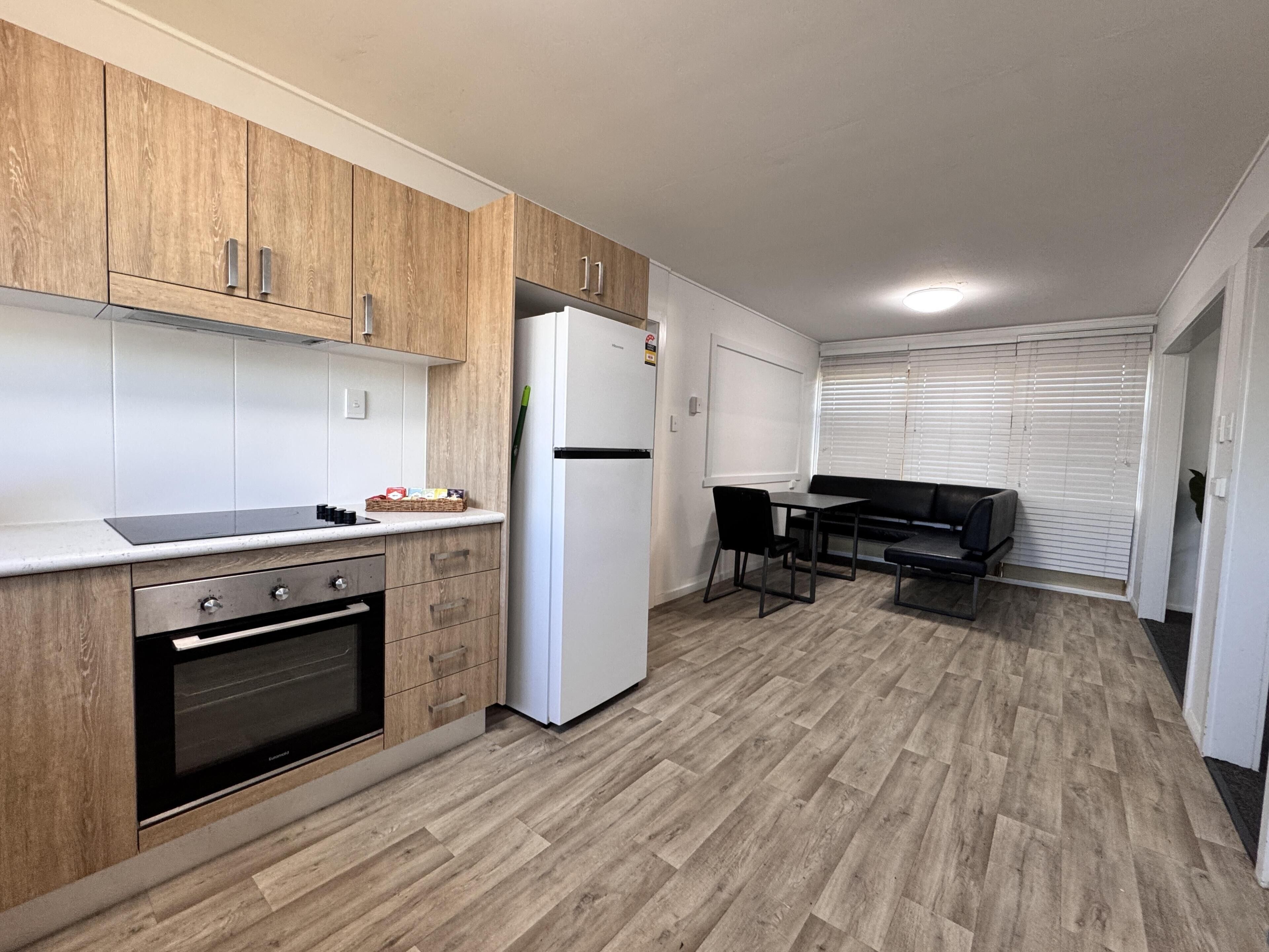 Een kleine keuken met houten kastjes, een inbouwoven, een witte koelkast en een eethoek met een zwarte tafel en stoelen.