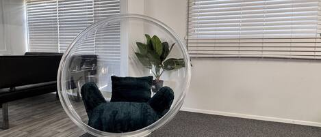 Una habitación con una silla de vidrio transparente, una planta en maceta, un banco y una ventana con persianas.
