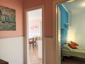 A room with a bed, a desk, and a chair. There is a painting on the wall and a door leading to another room.