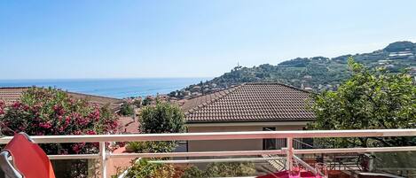 Un balcone con un tavolo apparecchiato per due, con vista su una cittadina costiera dai tetti in terracotta e un cielo azzurro intenso.