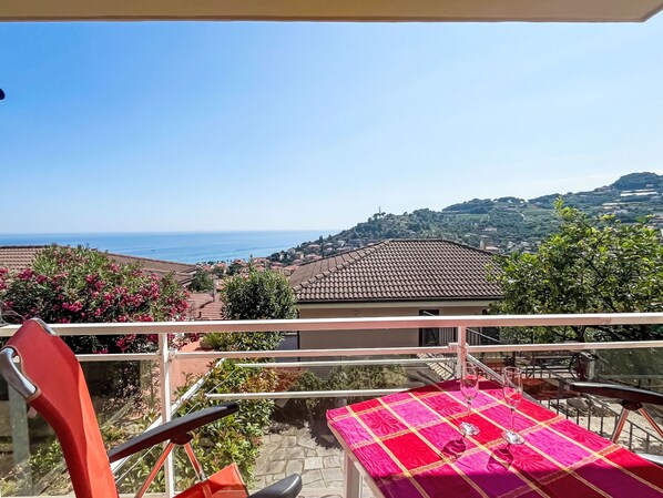 A balcony with a table set for two, overlooking a coastal town with terracotta rooftops and a clear blue sky.
