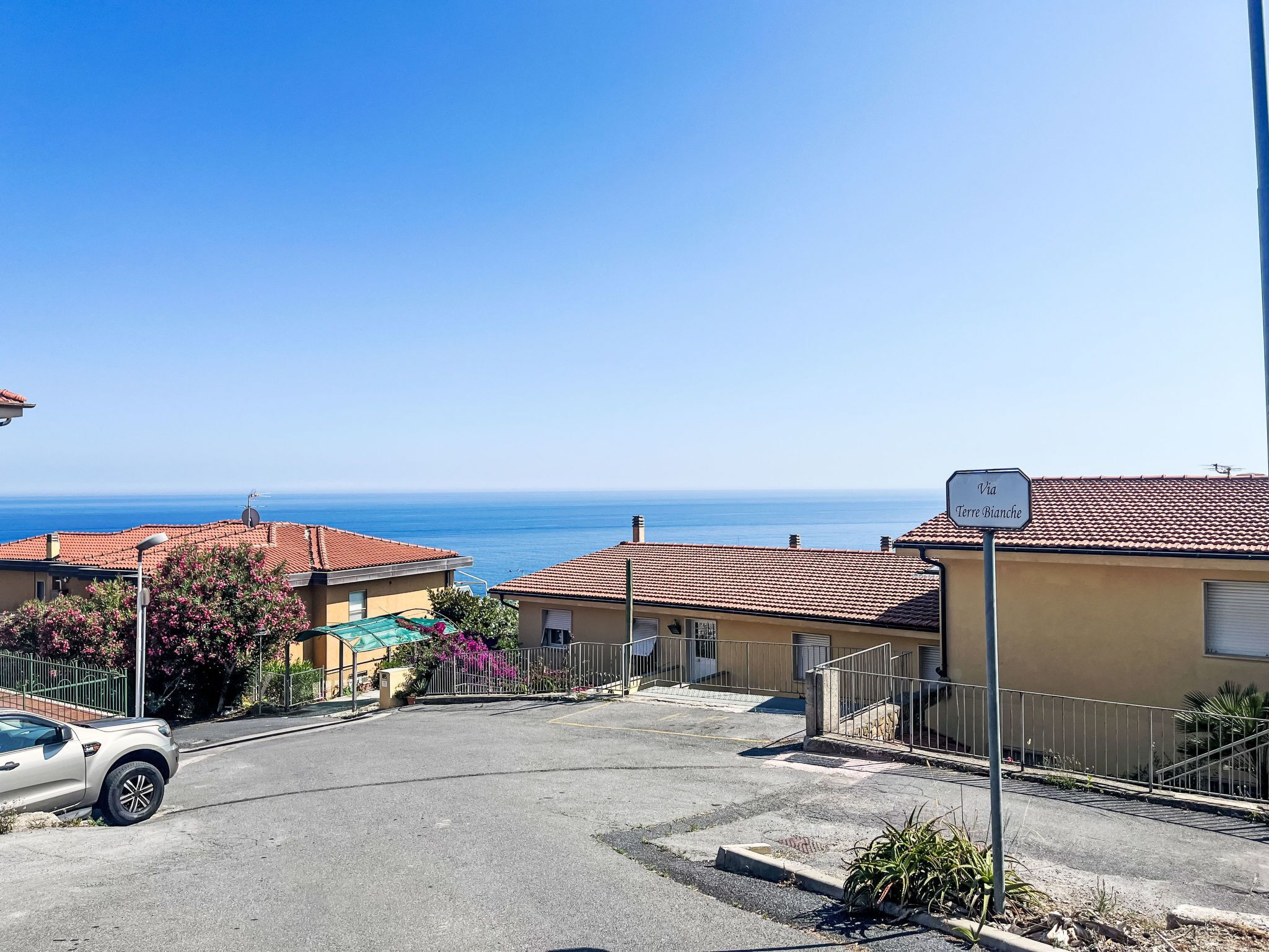 Eine Wohnstraße mit Häusern mit Terrakotta-Dächern, einem geparkten Auto und freiem Blick auf das Meer.