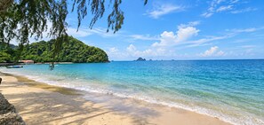 Ein Strand mit klarem türkisblauem Wasser, Sandküste und einer bewaldeten Hügelkette im Hintergrund.