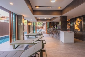 Un área de piscina interior moderna con sillones de descanso, un bar y vista a la piscina exterior.