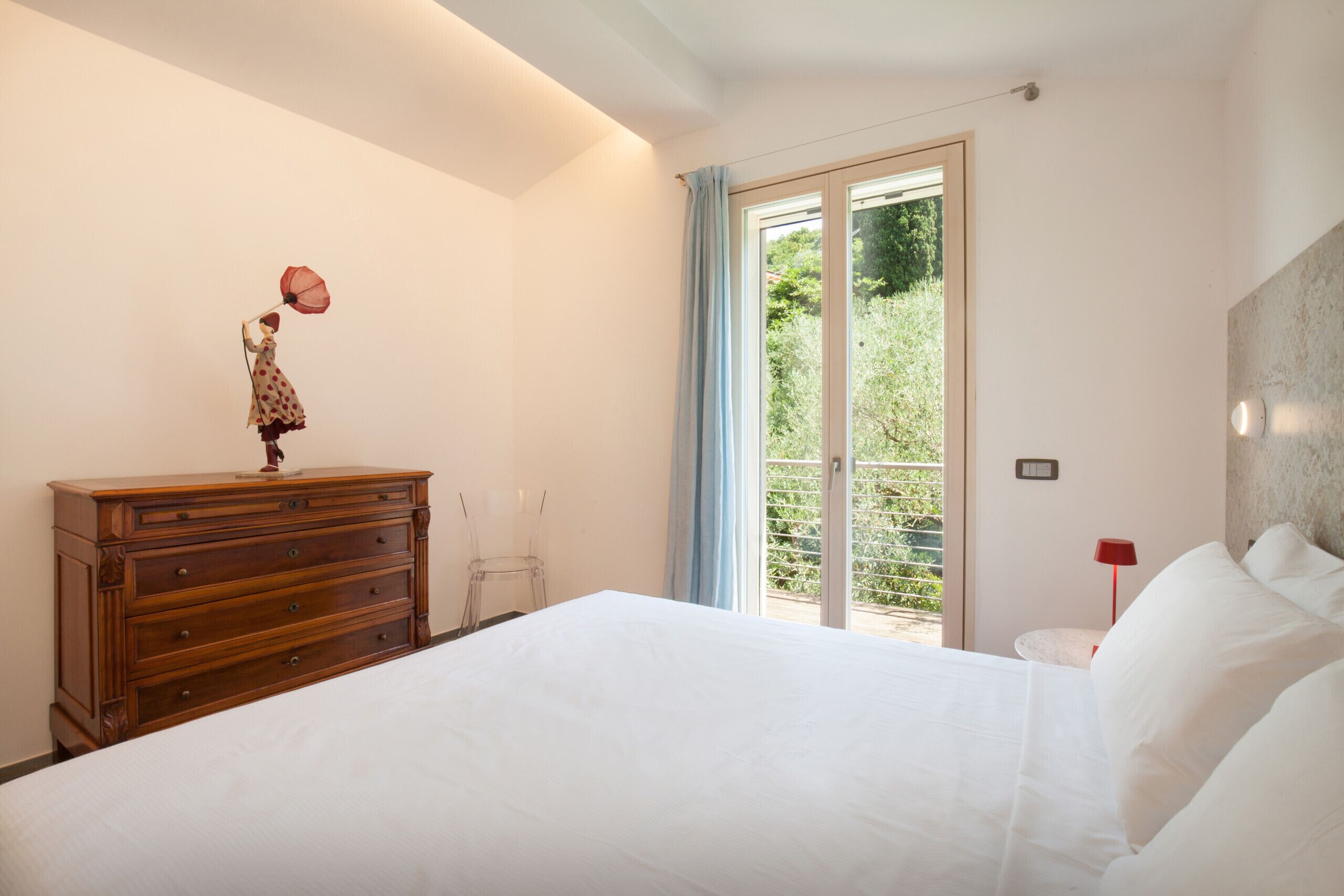 A bedroom with a bed, dresser, and a view of greenery through a sliding glass door.