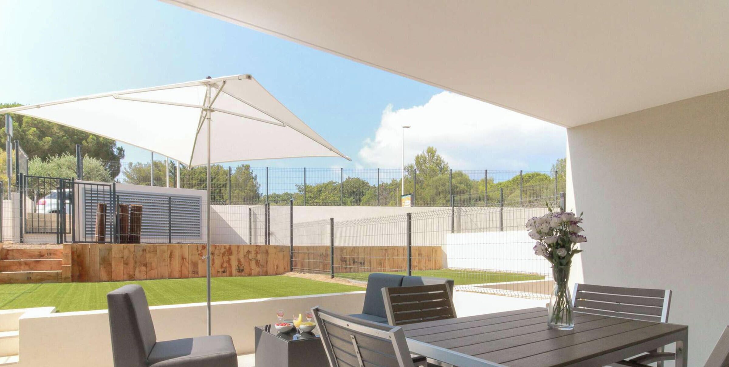 Un patio al aire libre con mesa y sillas, un paraguas grande y vistas a un área cercada con muelle de madera y césped.