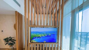 Un intérieur moderne avec un grand téléviseur à écran plat fixé sur un mur en lambris de bois, une plante en pot et de grandes fenêtres offrant une vue sur l’extérieur.