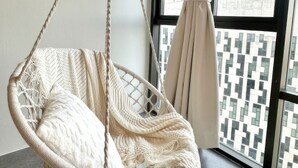 Una silla colgante de macramé con un cojín blanco en una habitación con un ventanal y vistas a edificios.