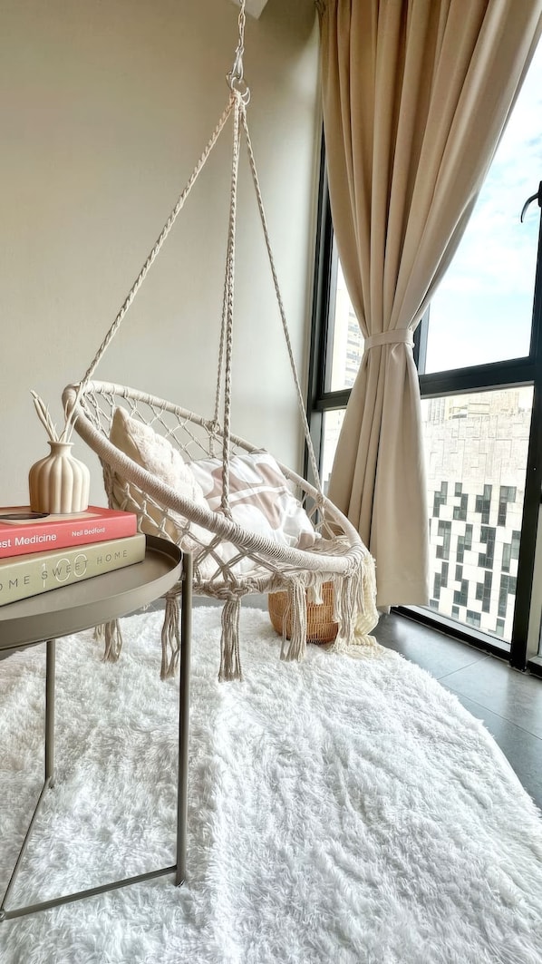 Una habitación con una silla colgante de macramé, un tapete blanco y mullido, una mesa con libros y vistas a edificios a través de amplios ventanales.