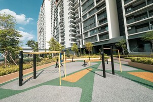Un parque infantil con columpios y una estructura para escalar, rodeado de vegetación y un edificio con varios balcones.