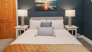 Una camera da letto con un letto, due comodini, un tavolino e un quadro raffigurante un tramonto sull'oceano.