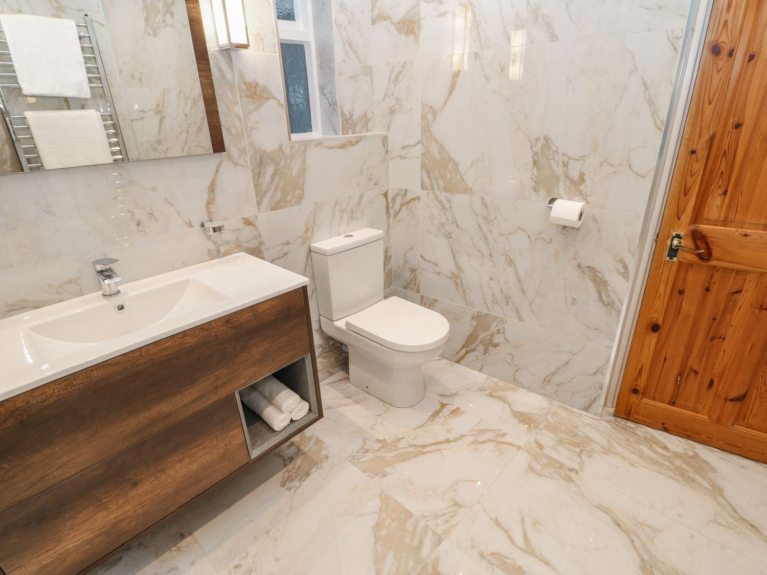 Un baño con suelo de mármol, tocador de madera, lavabo blanco, inodoro y puerta de madera.