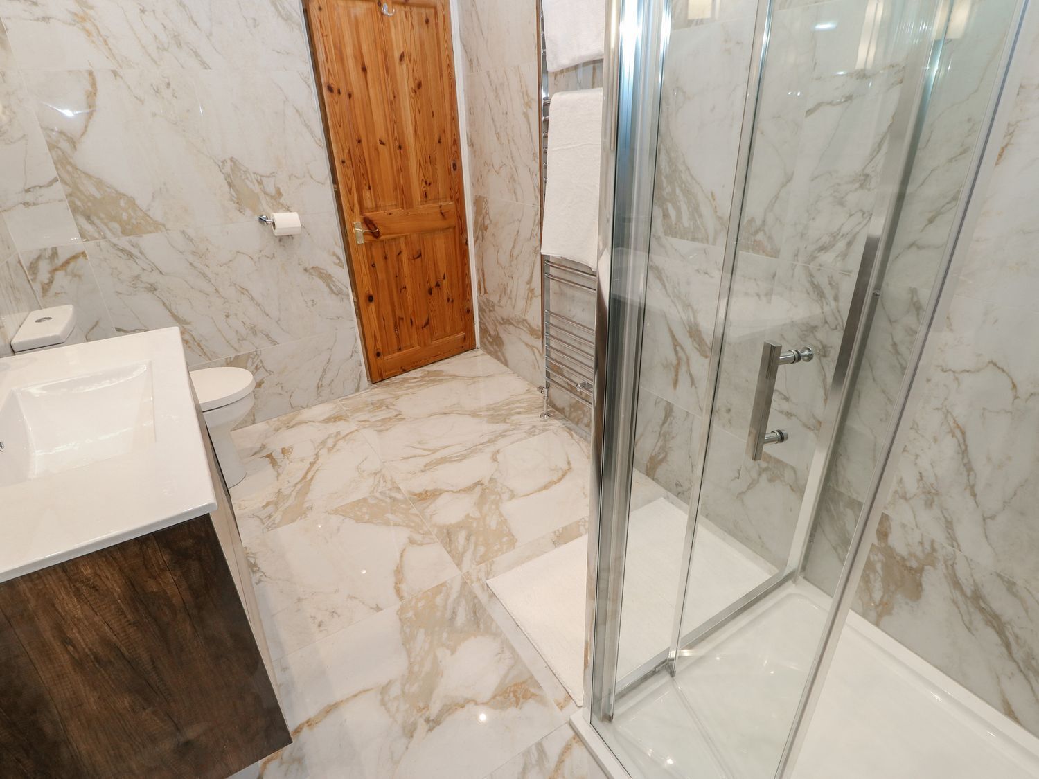 A bathroom with a marble floor, a wooden door, a glass shower enclosure, and a white sink.