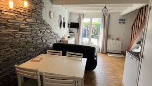 Ein Essbereich mit einer Steinwand, einem weißen Tisch und Stühlen. Es gibt ein schwarzes Sofa und einen an der Wand befestigten Fernseher.