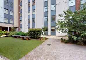 Un patio con zona verde, un sendero empedrado y un edificio de múltiples ventanales y fachada de ladrillo.