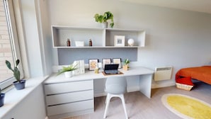 Une chambre moderne avec un bureau blanc, une chaise grise, un lit recouvert d’une couverture orange et une fenêtre agrémentée d’une plante.
