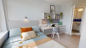 Une chambre moderne avec un lit, un bureau et une chaise. On y trouve une étagère avec des plantes et un cadre photo.