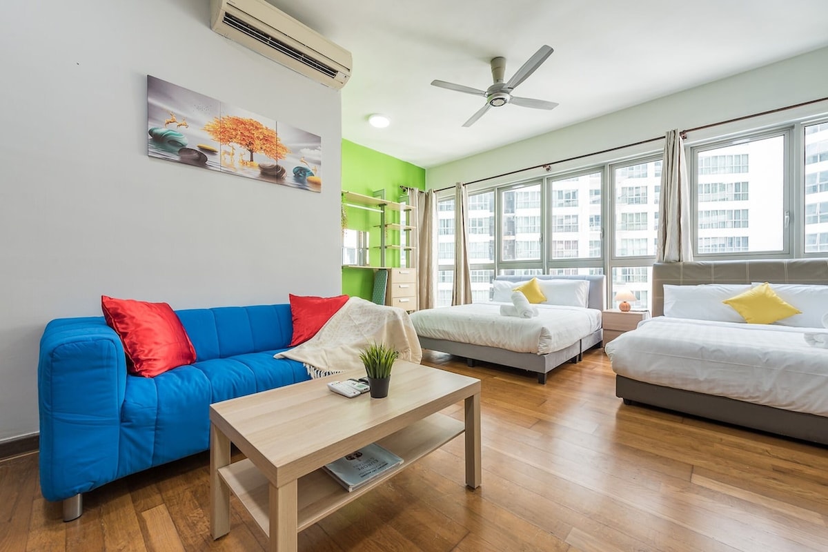 A modern hotel room with a blue sofa, a white bed, a wooden coffee table, and a large window offering a city view.