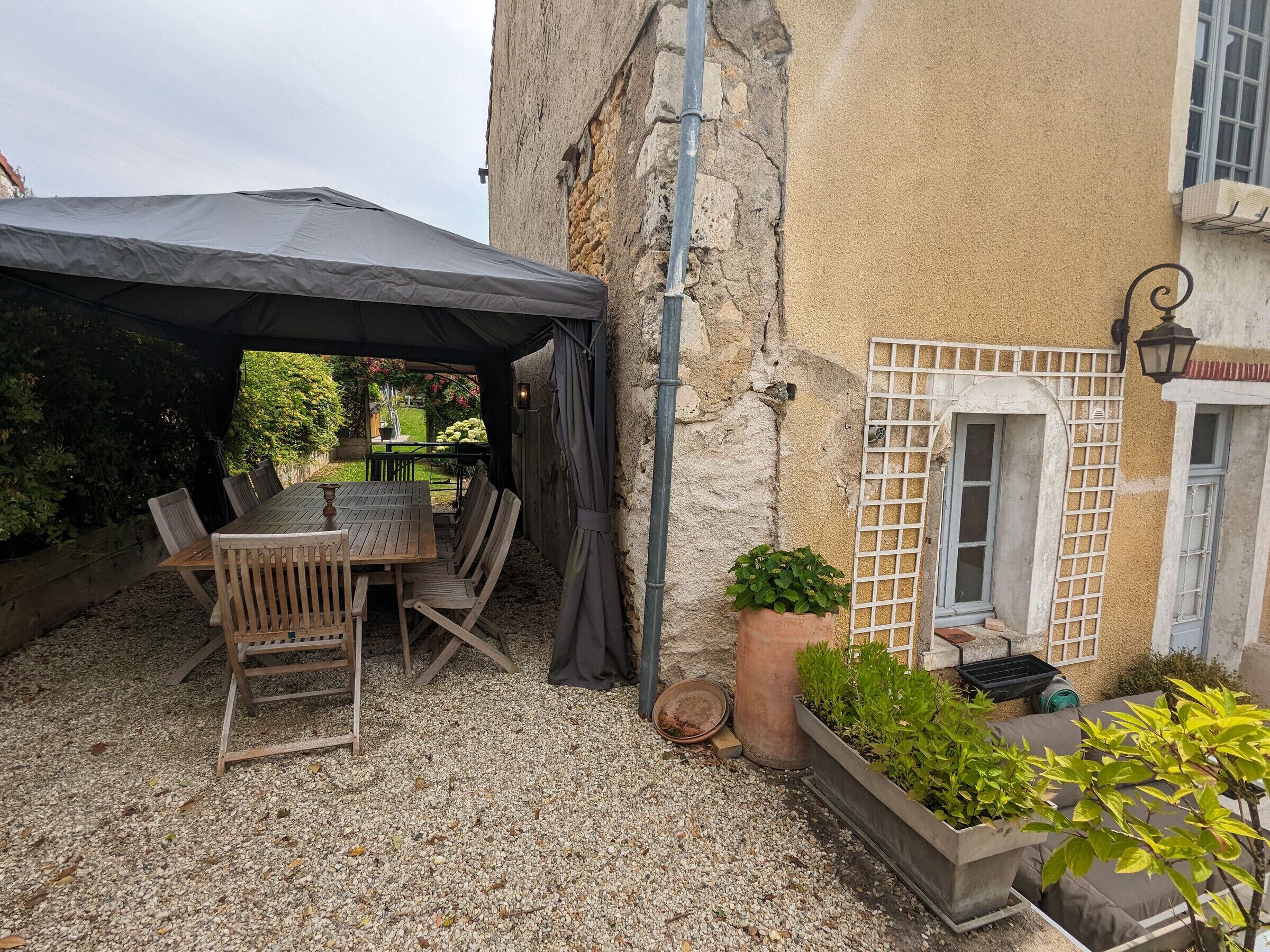 Eine Terrasse im Freien mit Holztisch und -stühlen, einem überdachten Bereich und Pflanzkübeln.