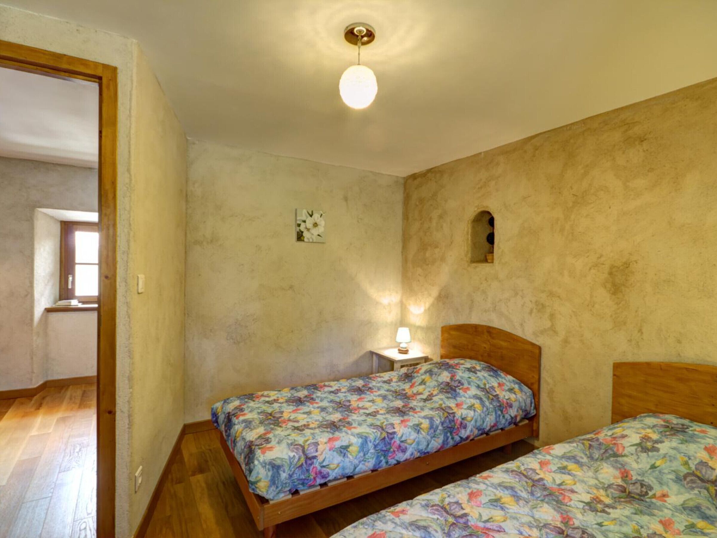 Une petite chambre à coucher avec deux lits en bois, une table de chevet, une lampe fixée au mur et une fenêtre.