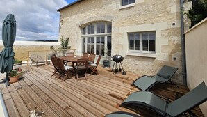 Een houten terras met een eettafel en stoelen, een barbecue en lounge fauteuils.