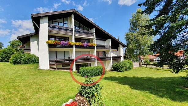 Un edificio moderno de dos pisos con balcones, un césped cuidado y abundante vegetación circundante.