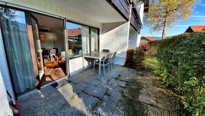 Un patio exterior moderno con una mesa y sillas, un vidrio que da a un espacio interior, y un jardín con arbustos y un árbol.