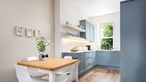 Een moderne keuken met een houten tafel en witte stoelen, blauwe kastjes en een raam met uitzicht op groen.