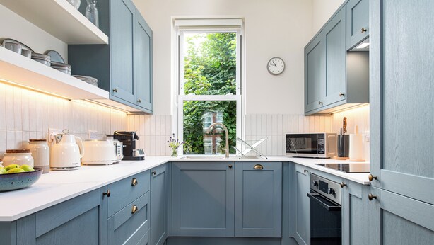 Een keuken met lichtblauwe kastjes, een wit aanrechtblad en een raam met uitzicht op groen.