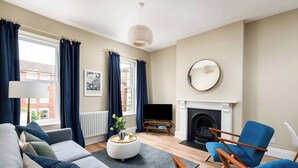 Een woonkamer met een bank, fauteuils, een open haard, een televisie en een raam met gordijnen.