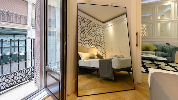 A modern bedroom with a large mirror, a bed with pillows, a nightstand, and a view of the outside balcony.